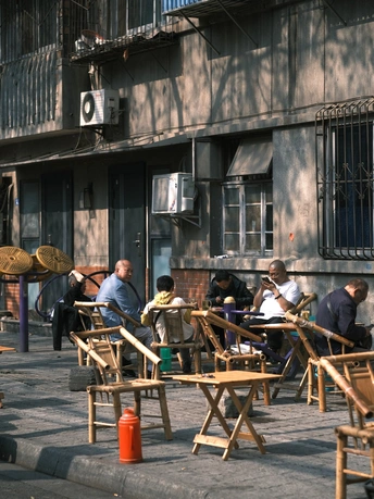 许多人坐在带有遮阳伞的桌子旁 位于一栋建筑旁边的人行道上。