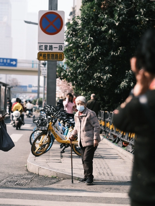 一个戴口罩的老先生站在人行道上 旁边有一辆自行车 行人从繁忙的街道上走过。