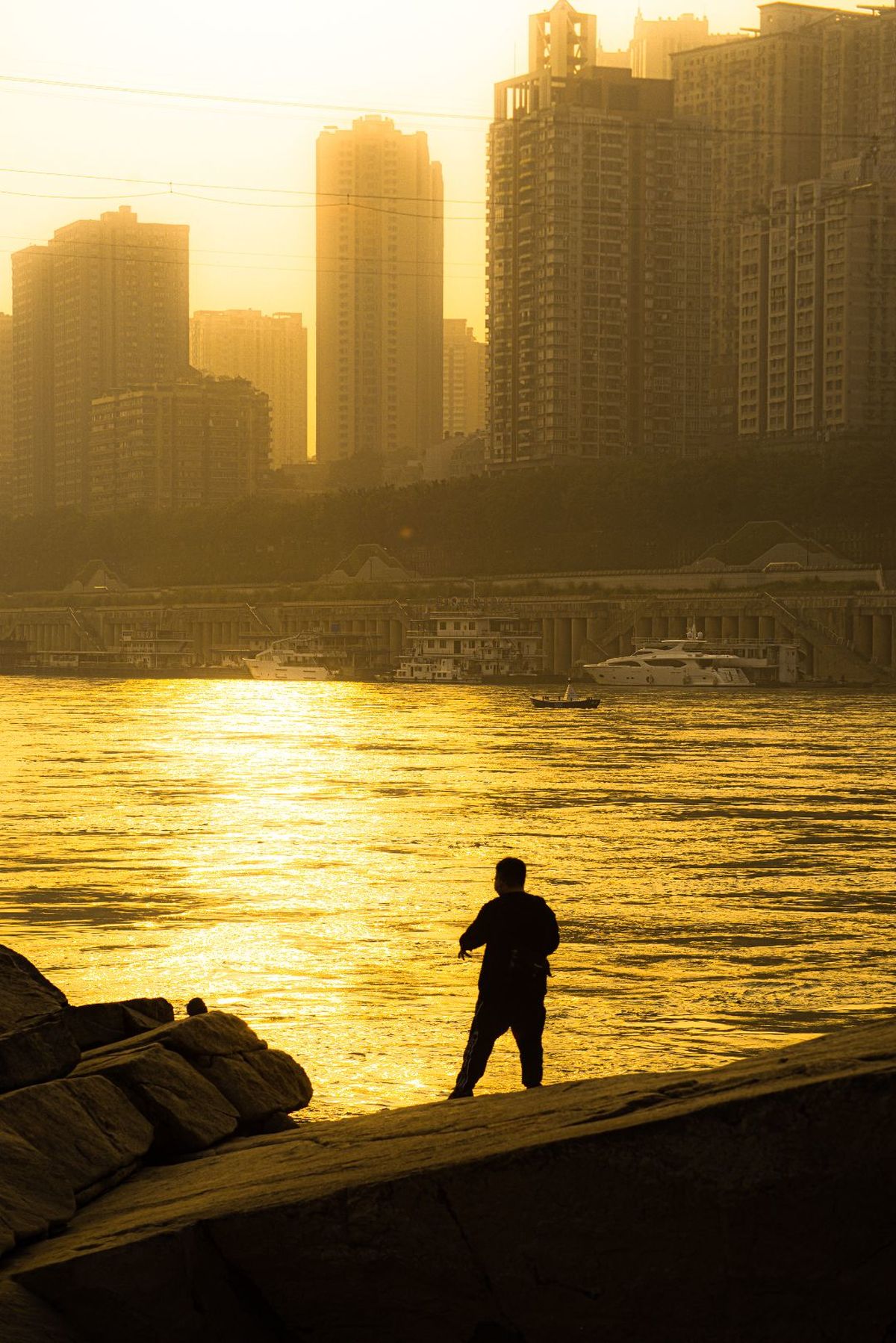 一个人沿着海滩走 背景是夕阳下的城市、水域和建筑物。