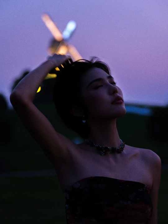 一个穿着连衣裙的年轻女子站在黄昏或夜晚的田野中 背景中有风车。