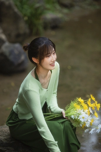 一个穿着绿色连衣裙的年轻女子拿着一束黄色的花坐在小溪边的岩石上