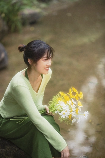 一个穿着绿色连衣裙的年轻女子拿着一束花坐在小溪里的岩石上