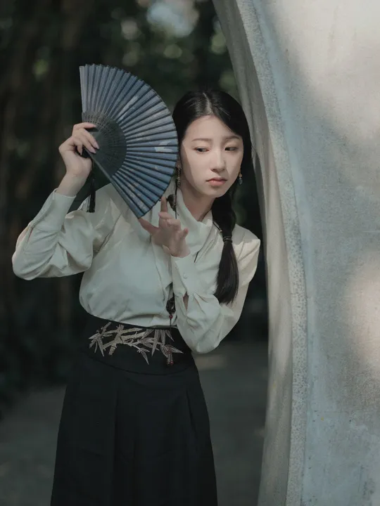 一位年轻女子手持扇子和雨伞站在一座雕像旁边