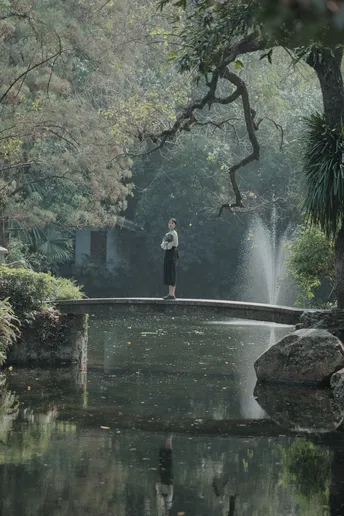 一个人站在公园里的桥上 周围有树木和水。