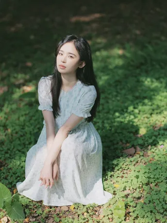 一个穿着白裙子的年轻女子坐在树下草地上