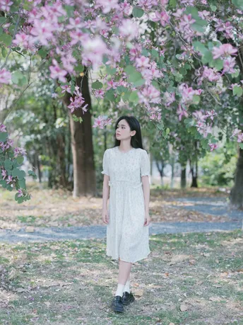 一位穿着白色连衣裙的年轻女子站在一棵开满粉色花朵的树下