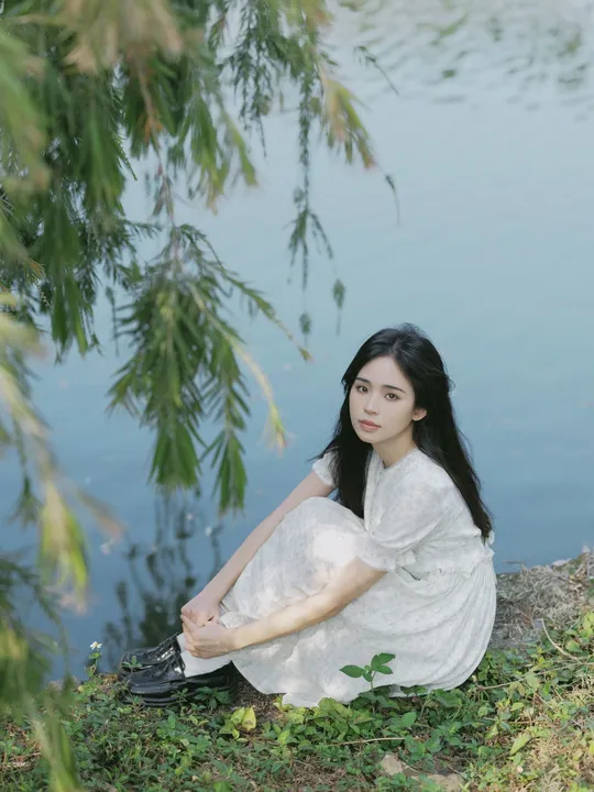 年轻美丽的女子穿着白色裙子坐在池塘或河边的岸边
