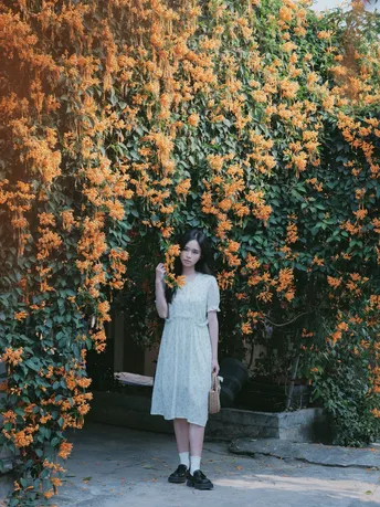 一位穿着白色连衣裙的年轻女子站在一堵长满橙色花朵的藤蔓墙前