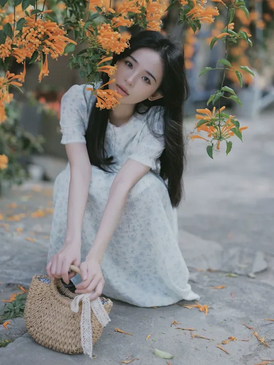 一位穿着白色连衣裙的年轻女子抱着一个包 坐在有橙色花朵的树下