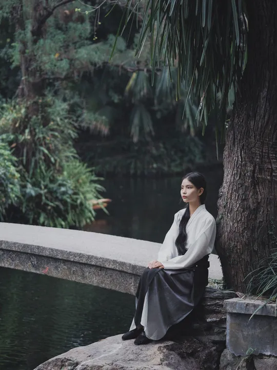 一位年轻女子坐在河边或湖边的一块岩石上 背景中有树木和桥。