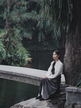 一位年轻女子坐在河边或湖边的一块岩石上 背景中有树木和桥。