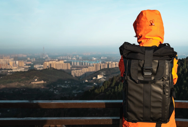 一个穿橙色夹克背有背包的男人坐在山顶的 bench 上 俯瞰城市。