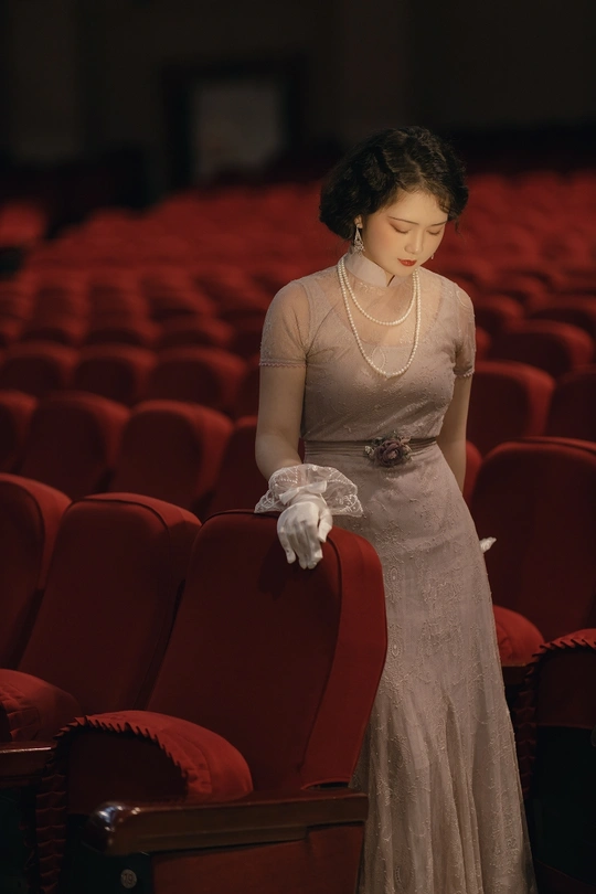 一位穿着白色连衣裙和白色手套的女人站在一个带有红色椅子的音乐厅里。