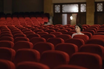 一个年轻女子独自坐在空荡荡的剧院里 有红色的座位 后面有屏幕。