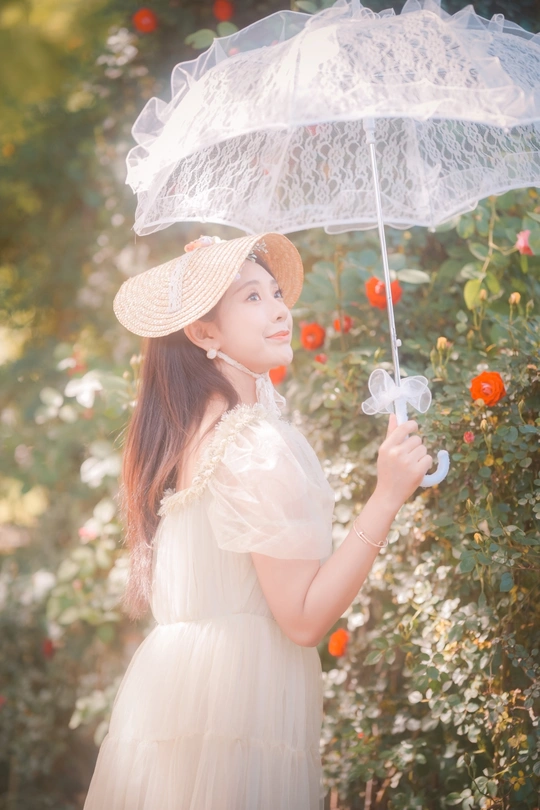 一位穿着白色连衣裙和帽子 手持透明雨伞的年轻女子在花园里。