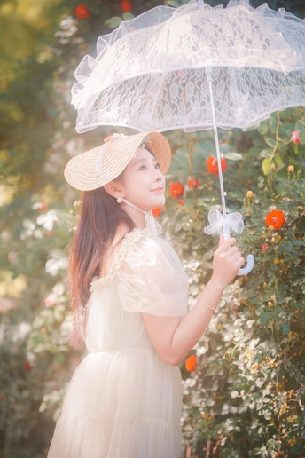 一位穿着白色连衣裙和帽子 手持透明雨伞的年轻女子在花园里。