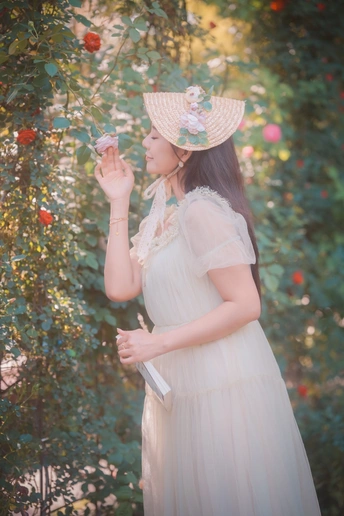 一位美丽的年轻女子穿着一件白色连衣裙和一顶草帽 手里拿着一朵花站在花园里。