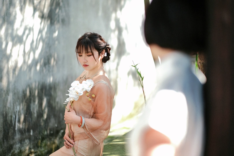 一位身穿白色连衣裙、手持鲜花的年轻女子站在瀑布和镜子前