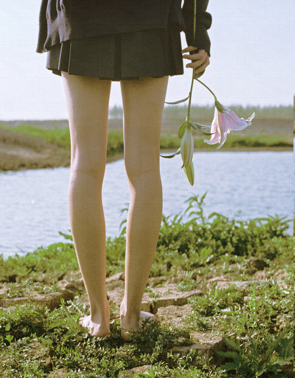 一位穿着连衣裙的年轻女子手持一束花和一把伞 站在湖边或水体旁边。