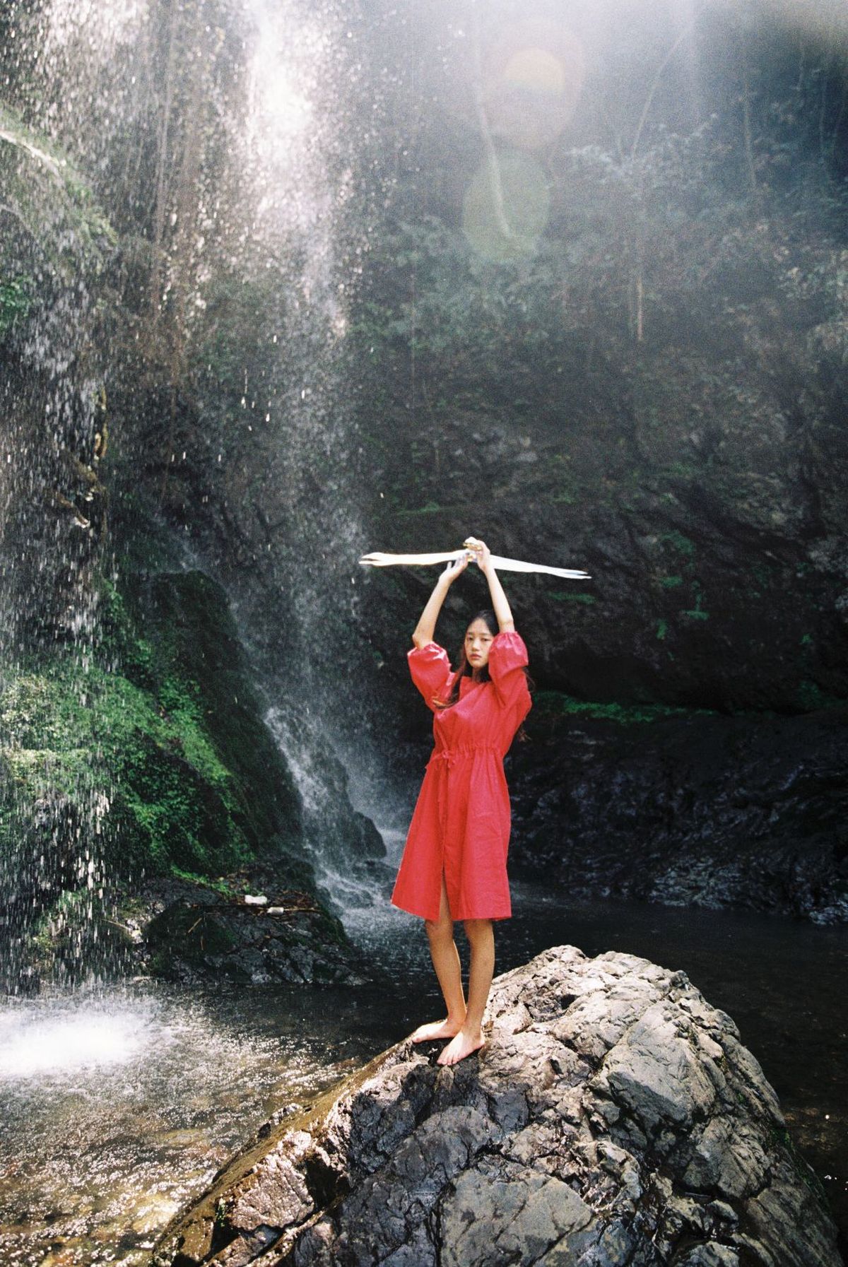 一位穿着红色连衣裙的女孩举着棍子站在瀑布前的岩石上