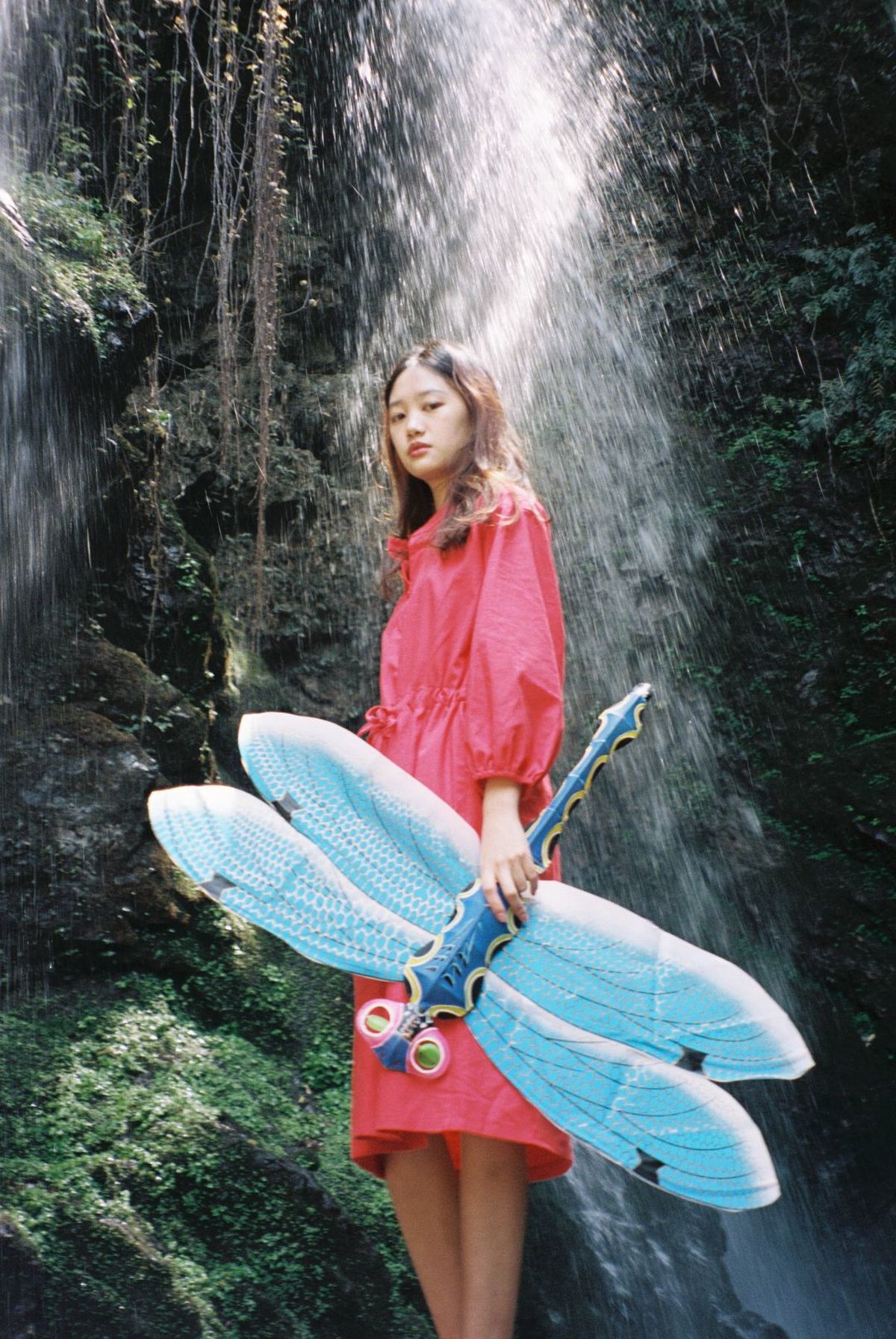一位穿着红色连衣裙和雨衣 手持冲浪板站在瀑布前的女人