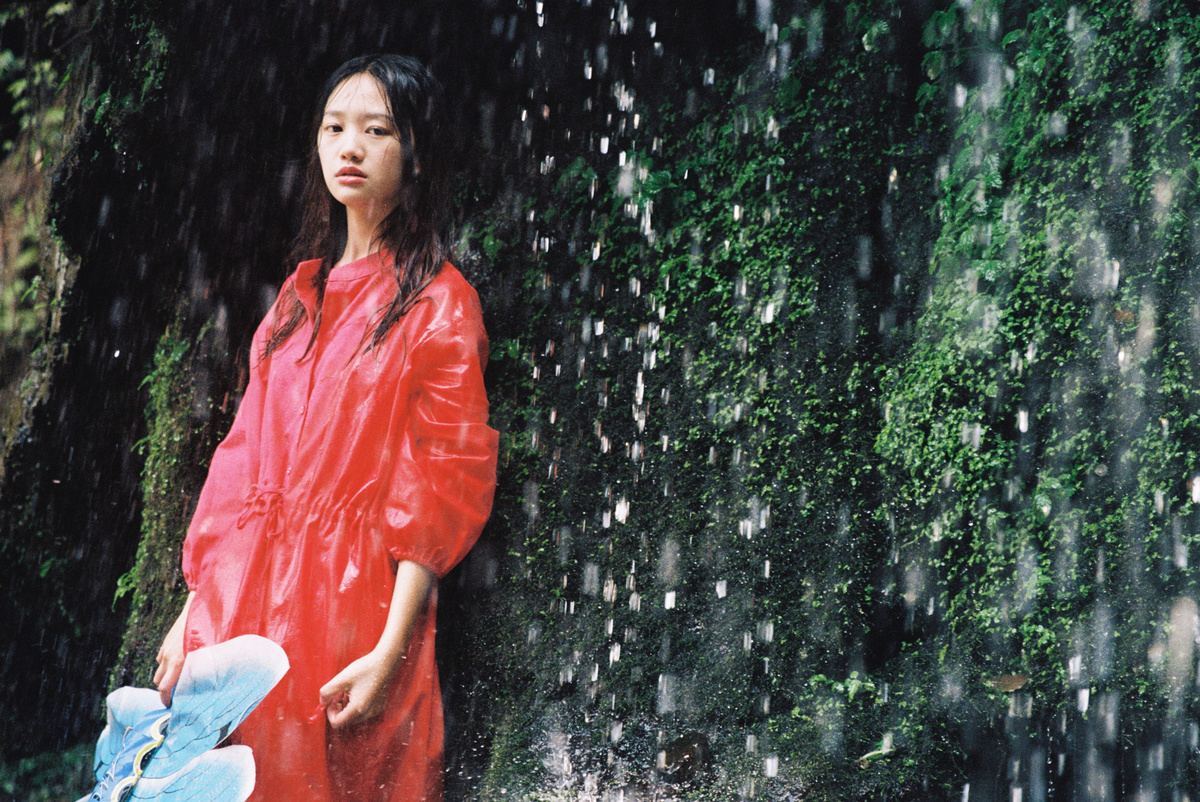 一位穿着红色连衣裙和拿着雨伞的年轻女子站在瀑布下的水帘中 手里抱着一只泰迪熊。