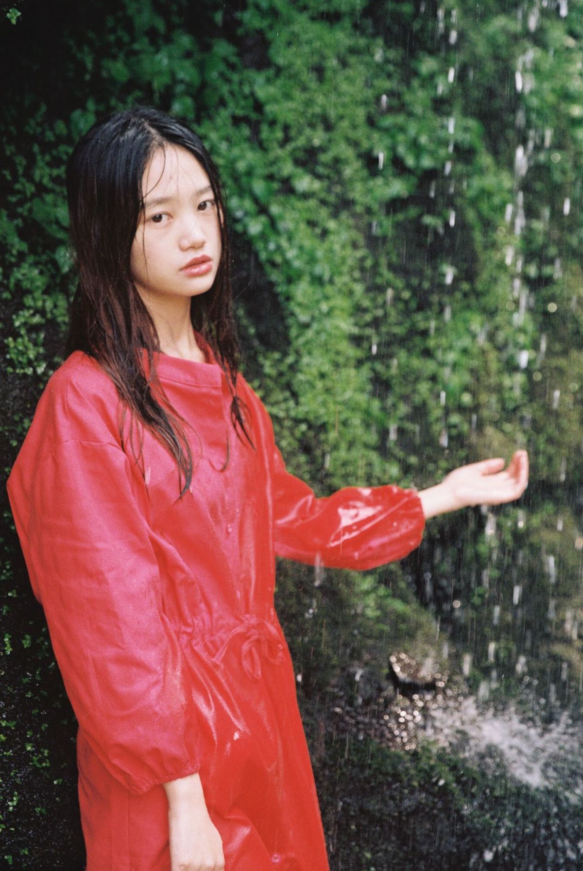 一位穿着红色雨衣和红色连衣裙的女士站在瀑布下 冒着雨 打着红色雨伞。