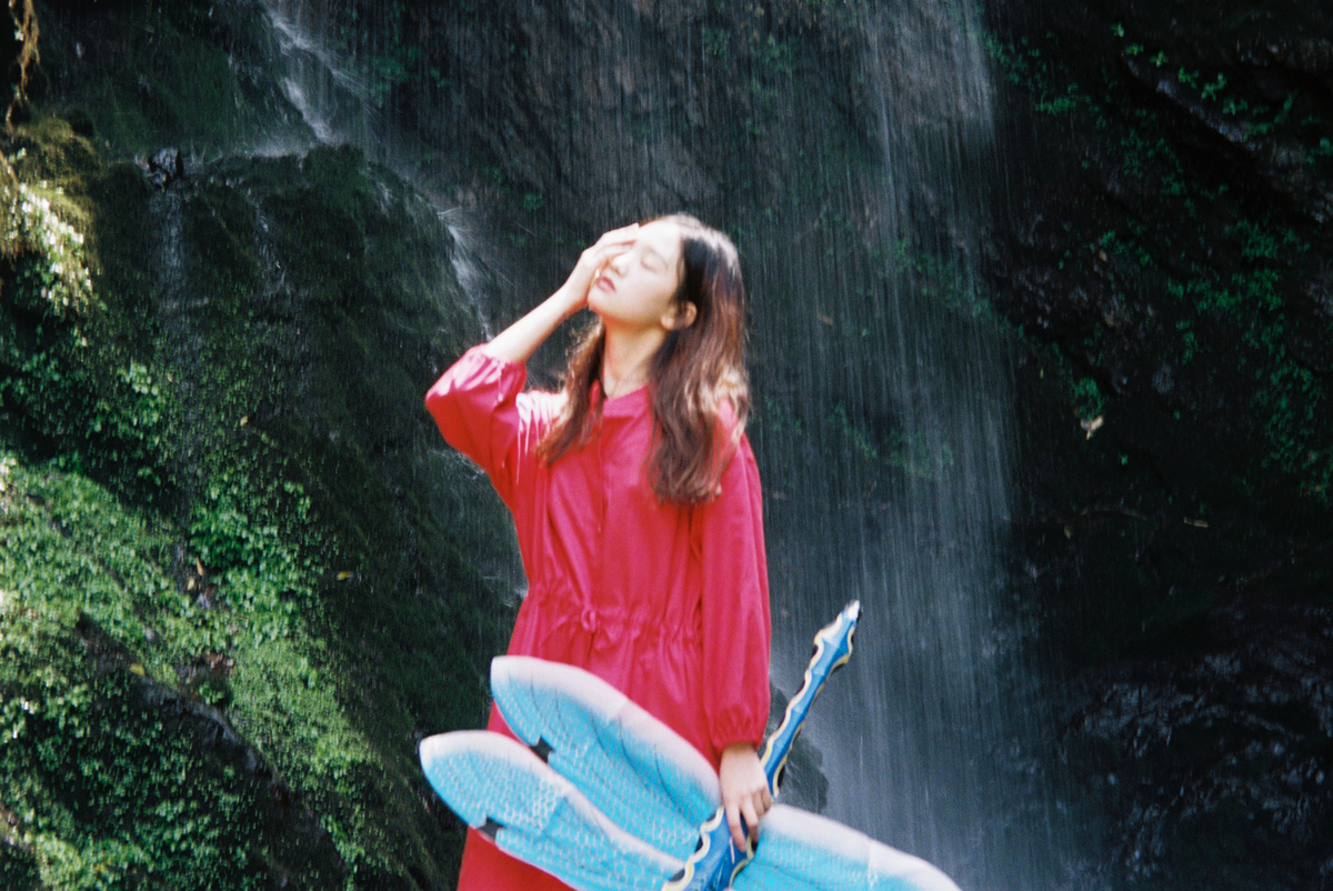 一位穿着红裙子的年轻女子站在瀑布前 手里拿着一块蓝色的冲浪板。