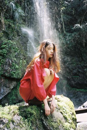 一位穿着红色连衣裙和雨衣的年轻女子坐在瀑布前的岩石上。