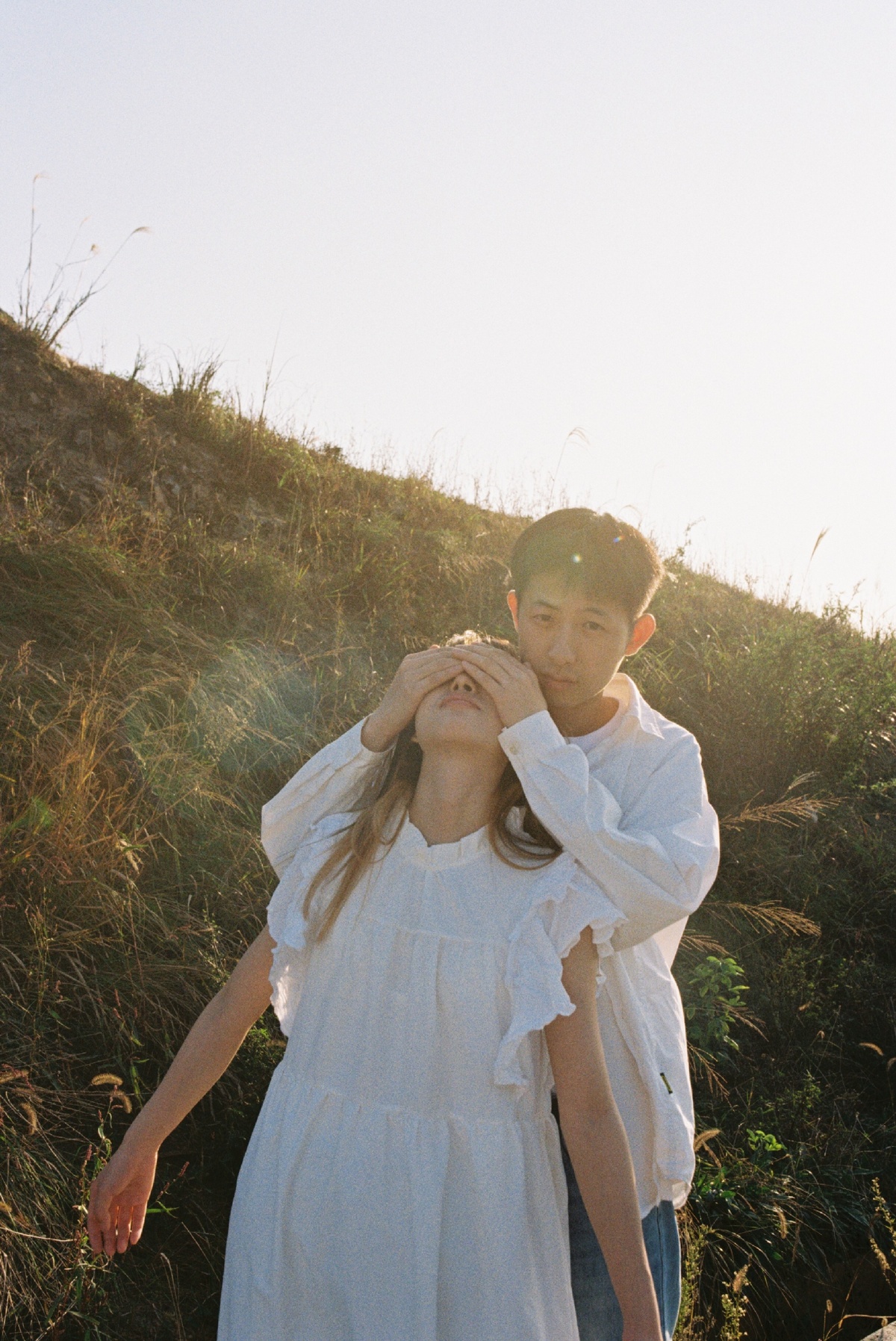 一个穿着白裙子的年轻女孩站在一个田野里 双手捂住眼睛 一个年轻男孩沿着山坡走上来。