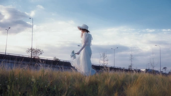 一位穿着白色连衣裙和帽子 手里拿着一朵花站在草地上的年轻女子。