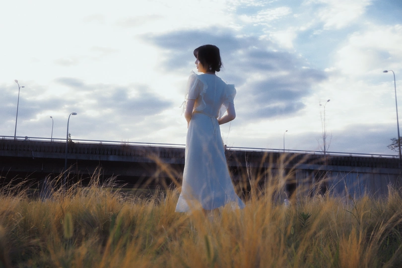 一位穿着白色连衣裙的女士站在草地上 背景中有一座桥。