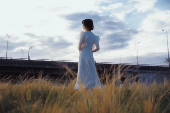 一位穿着白色连衣裙的女士站在草地上 背景中有一座桥。
