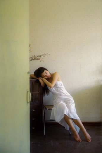 一个穿着白色连衣裙的年轻女子坐在椅子上 倚靠在房间角落的墙上。