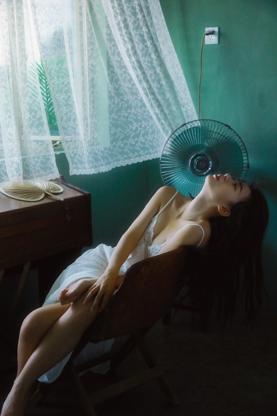 一个年轻女子躺在有风扇、窗户和一张有椅子的床上的房间里