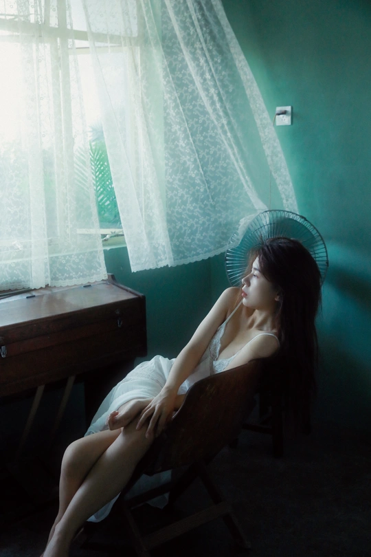 一个年轻女子坐在一个黑暗的卧室里的椅子上 有风扇和窗户。