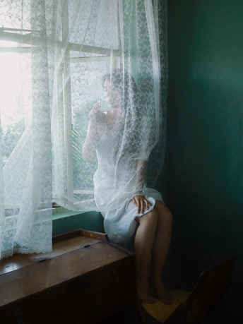 一个女孩坐在房间里 透过白色的窗帘看着窗外。