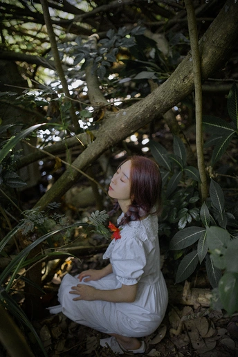 一个穿着白色连衣裙的年轻女子坐在树木和灌木丛中。