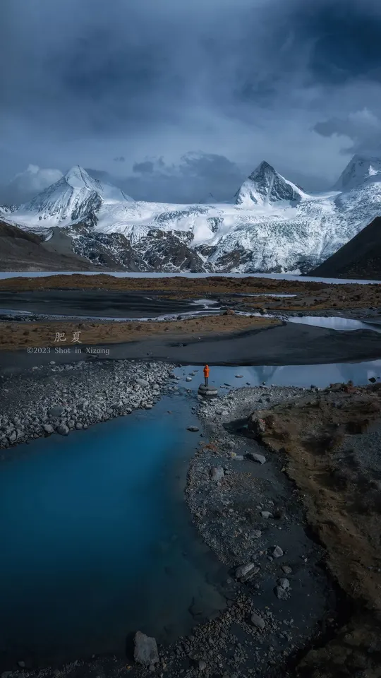 一个人站在一个小湖边 湖里有蓝色的水 前面是雪山。