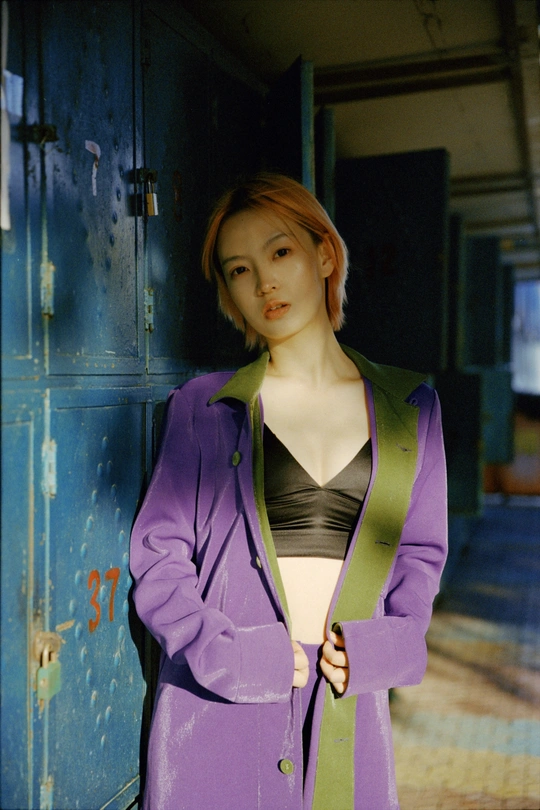 一位穿着紫色外套的女子站在一个废弃的更衣室旁边 旁边有一个绿色的人形模特。