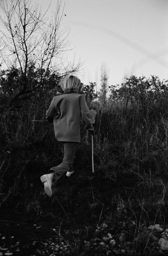 一个黑白照片 一个年轻男孩穿过茂密的草丛和灌木丛 前景是木棍 背景是树木。