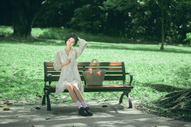 一个穿着白色连衣裙的年轻女子坐在公园的长椅上 手里拿着一个篮子。