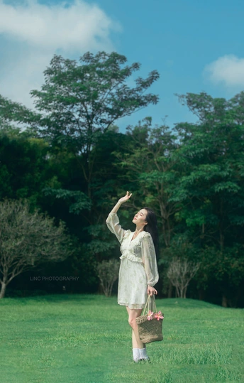 一位穿着白色连衣裙的年轻女子手提篮子和雨伞穿越草地 而另一位穿着白色连衣裙的女孩手里拿着一个袋子。