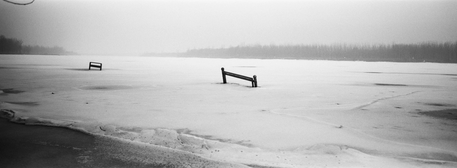 一张黑白照片 展示了一个空荡荡的长椅和一张雪地上的公园长椅 周围有冰和水。