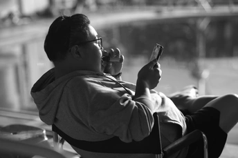 一张黑白照片 一个男子坐在椅子上 看着他的手机 给窗户拍照。