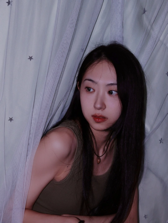 一个年轻女子坐在窗边一张床的床单上 窗帘垂下