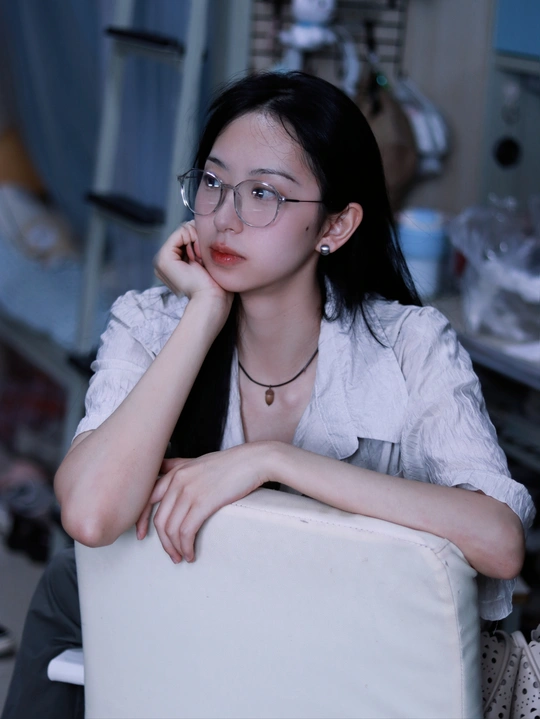 一个戴眼镜的年轻女子坐在一个凌乱的房间里的一把白色椅子上