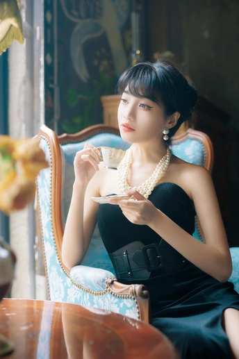 一位身穿黑色连衣裙、佩戴珍珠首饰的年轻女子坐在桌子旁的椅子上喝茶