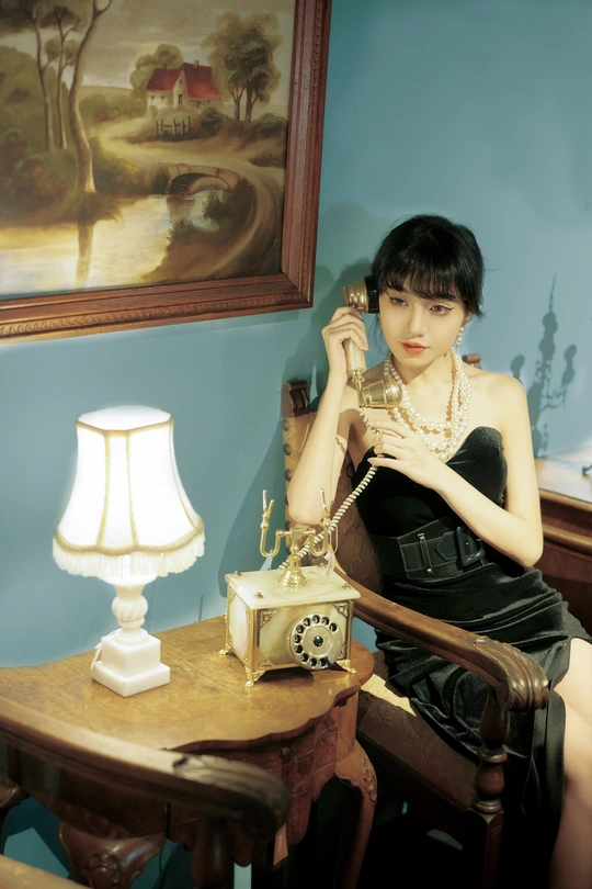 一个穿着黑色连衣裙的年轻女子坐在桌子旁的椅子上 桌子上有一部旧电话和一盏灯