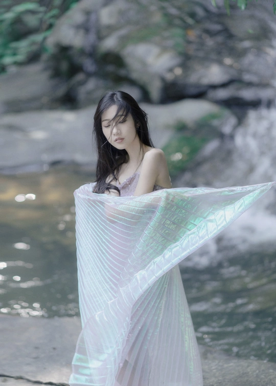 一位身穿白色连衣裙、披着粉色披肩的美丽年轻女子站在溪流中